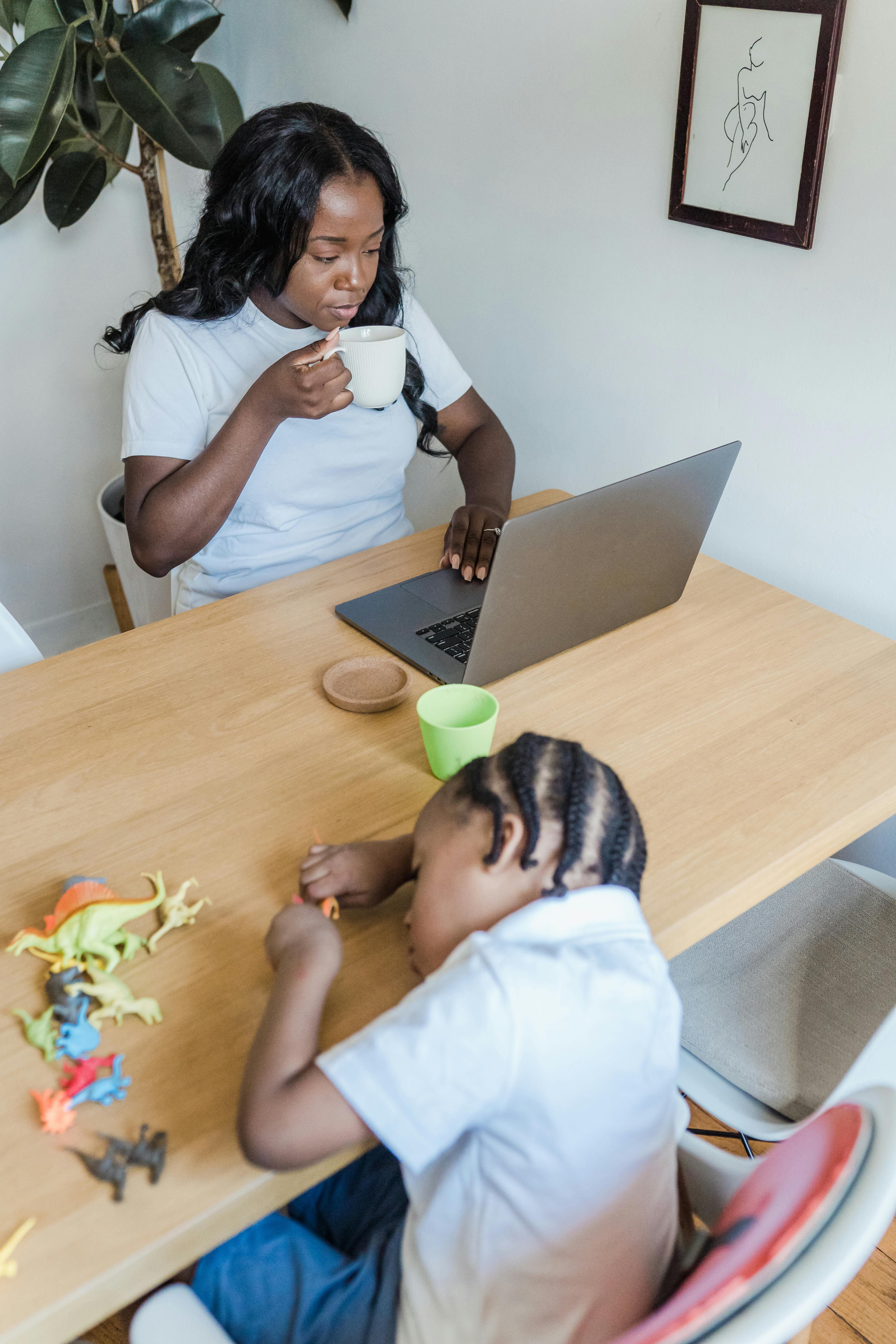 A woman sipping coffee while working on a laptop as her child plays with toys at home.