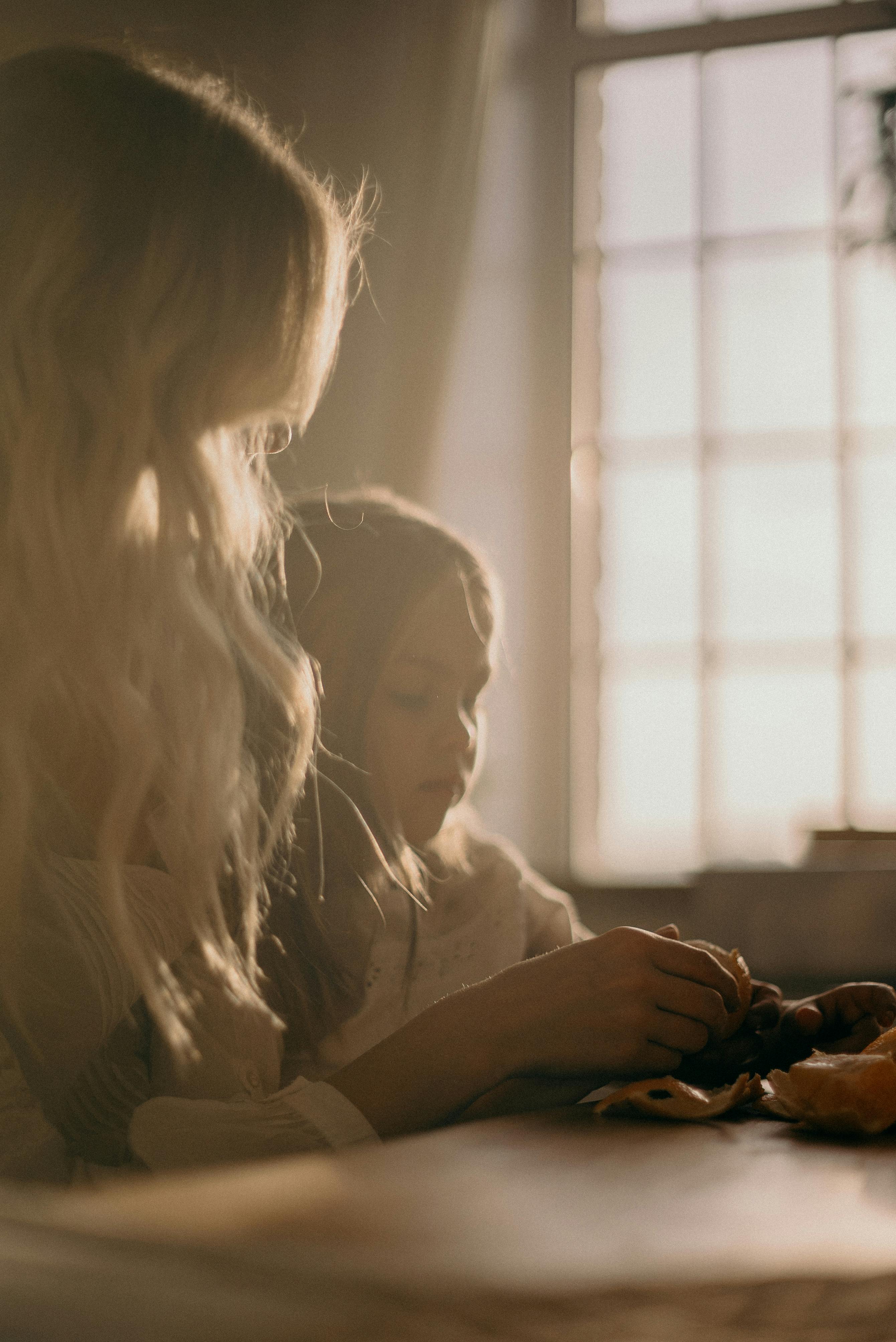 A warm, intimate moment between a mother and daughter sitting by a sunlit window indoors.